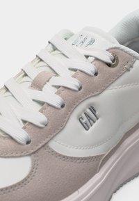 Άσπρο και μπεζ αθλητικό παπούτσι με λευκά κορδόνια, σουέτ λεπτομέρειες και το λογότυπο "GAP" στο πλάι και στη γλώσσα.