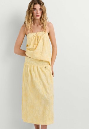 Jonge vrouw met golvend haar, gekleed in een gele strapless top en bijpassende midi-rok met bloemenborduursel, staand tegen een witte muur.