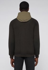 Mörkgrön stickad tröja med huva i olivgrön. Har ribbad textur och långa ärmar. Bakifrån syns kontrasterande tyg och design.