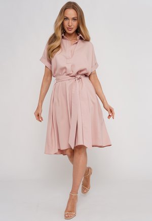Kobieta spaceruje w jasnoróżowej sukience do kolan z krótkimi rękawami, kołnierzykiem i paskiem do wiązania, w zestawieniu z beżowymi sandałkami na obcasie z paskiem wokół kostki.