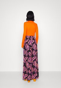 Kvinde med ryggen til iført en skarp orange langærmet top, sorte bukser med vidde med mønster af pink og lilla blade, samt orange blokhæle.