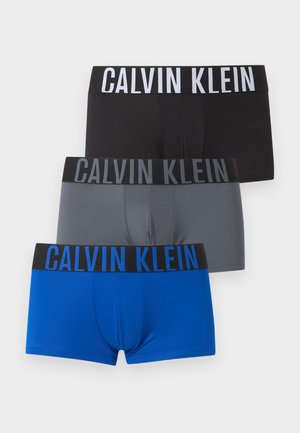 Mies, jolla on lyhyet tummat hiukset ja mustat Calvin Klein -bokserit, seisoo paljain jaloin valkoista taustaa vasten.