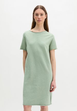 Kobieta z długimi brązowymi włosami, ubrana w luźną, krótko-rękawową, jasnozieloną sukienkę sięgającą kolan na jednolitym białym tle.