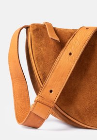 Tan suede crossbody bag with a curved shape and a smooth leather strap, featuring visible stitching and a buckle attachment detail.
