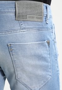 Ljust blå denimjeans med en texturerad grå midjebandspatch, kontrasterande sömmar och silverfärgad hårdvara på bakfickorna.