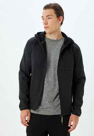 Sort softshell-jakke med hætte, forlommer, lynlås og en glat tekstur, båret over en grå T-shirt og sorte bukser.