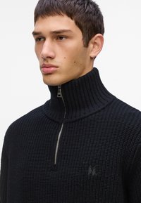 Pulover negru din tricot cu guler înalt, cu fermoar pe jumătate, având un detaliu subtil cu logo și un material texturat.