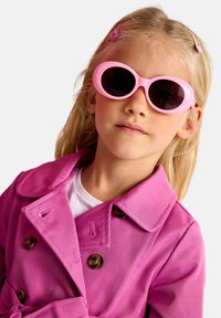 Trench coat rosa con bottoni marroni; abbinato a occhiali da sole ovali rosa con lenti scure; capelli biondi con fermagli coordinati.