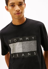 Camiseta de algodón negra con un logotipo rectangular gris que muestra el texto "TOMMY JEANS", cuello redondo y mangas cortas. Diseño simple y moderno.