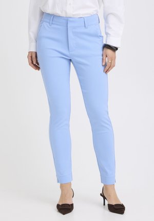 Kobieta ubrana w dopasowane, jasnoniebieskie spodnie sięgające kostek, w parze z czarnymi szpilkami z noskiem w szpic oraz białą koszulą z długim rękawem.