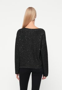 Maglione nero con una vestibilità ampia, tessuto testurizzato con glitter argentati, scollatura ampia e orlo e polsini a coste. Indossato sopra pantaloni neri.