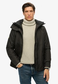 Zwarte geïsoleerde jas met een capuchon, ritssluiting en zijzakken, gedragen boven een lichtgrijze gebreide turtlenecktrui en blauwe spijkerbroek.