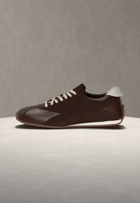 Καφέ sneaker χαμηλού προφίλ με λευκά κορδόνια και λευκή ταμπέλα στη φτέρνα, εμφανίζεται σε ουδέτερη επιφάνεια και φόντο.