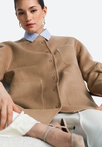 Bruine fleece jas met knopen en grote zakken, gedragen over een lichtblauw gestreept overhemd met kraag. Model draagt beige schoenen.