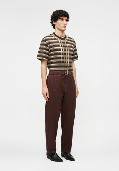 Νεαρός άνδρας όρθιος, φοράει καφέ ριγέ κοντομάνικο πουκάμισο, καφέ παντελόνι με υφή και μαύρα μυτερά παπούτσια, μπροστά σε ένα απλό φόντο.