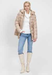 Giacca puffer beige con cappuccio, motivo trapuntato, maglione bianco a maglia, jeans azzurri chiari e stivali alti beige lucidi.