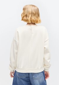 Personne aux cheveux blonds courts et ondulés portant un haut blanc ample à manches longues et un jean bleu en denim, tournée dos à la caméra.