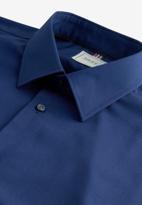 Chemise bleue marine avec un col pointu et un seul bouton visible, étalée à plat sur un fond blanc.