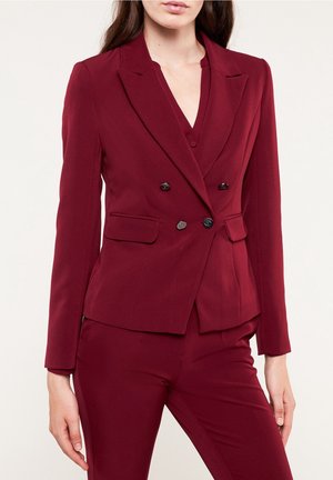Kvinde iført en tætsiddende bordeaux dobbeltradet blazer med matchende bukser mod en ensfarvet lys baggrund.