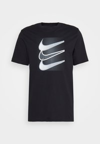 Svart kortärmad T-shirt med tre överlappande vita och grå Nike swoosh-logotyper på framsidan, mot en prickig rektangulär bakgrund.