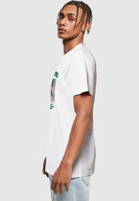 Hvid bomulds-t-shirt med korte ærmer, prydet med et grafisk print i grøn og en afslappet pasform. Modellen poserer sidelæns mod en neutral baggrund.