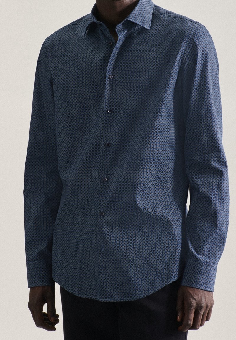 Man som bär en långärmad blå mönstrad skjorta med krage, stående mot en helt ljus bakgrund.