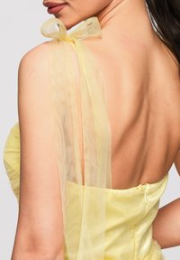 Gros plan du dos d'une femme portant une robe jaune sans bretelles avec un nœud jaune transparent noué sur son épaule.