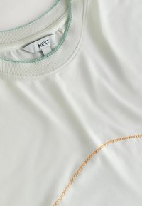 White crew neck shirt with green stitching along the collar and orange curved stitching accent on the fabric near the bottom right.