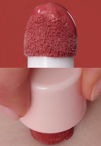 Applicatore tondo con punta morbida e testurizzata, rivestita da una tinta rossastra. Un cappuccio rosa tenue è tenuto sopra una superficie cutanea, mostrando l'uso.