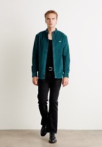 Camisa de botón de pana color verde azulado sobre una camiseta sin mangas negra, combinada con pantalones negros y zapatos negros, con un diseño simple y líneas limpias.