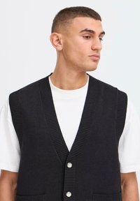 Jonge man met kort haar die een zwarte knoopvest over een wit T-shirt draagt, kijkend naar rechts tegen een eenvoudige lichte achtergrond.