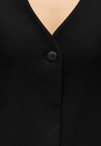 Blazer noir à col en V, doté d'une fermeture à un seul bouton noir et d'un tissu texturé, mettant en valeur un design élégant et minimaliste.