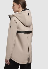 Beige vattentät jacka med en passformshuva, svarta detaljer och en slits bak. Har ett synligt logoband över ryggen.