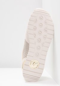 Semelle de sneaker rose clair avec une bande de roulement texturée, présentant un logo circulaire doré et des détails en mesh sur les côtés. Matière en caoutchouc, forme arrondie.