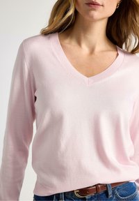 Lyserød V-hals sweater med lange ærmer, glat tekstur og ribkant ved halsen, kombineret med denimjeans og et brunt bælte.