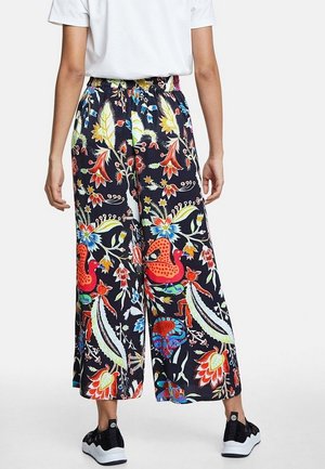 Persoon die zwarte wijde broek met kleurrijke bloemen- en dierenprint draagt, een wit T-shirt met korte mouwen en zwarte sneakers, en met de rug naar de camera staat.