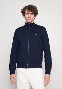 Sweatshirt zippé navy avec col montant, poignets et ourlet côtelés. Présente un petit logo vert Lacoste sur la poitrine gauche. Texture lisse.