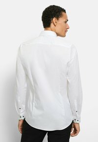 Weißes Langarmshirt mit figurbetontem Schnitt, klassischem Kragen und schwarzen Knopfakzenten an den Manschetten und Seitenähten. Glatte Textur, keine Muster.