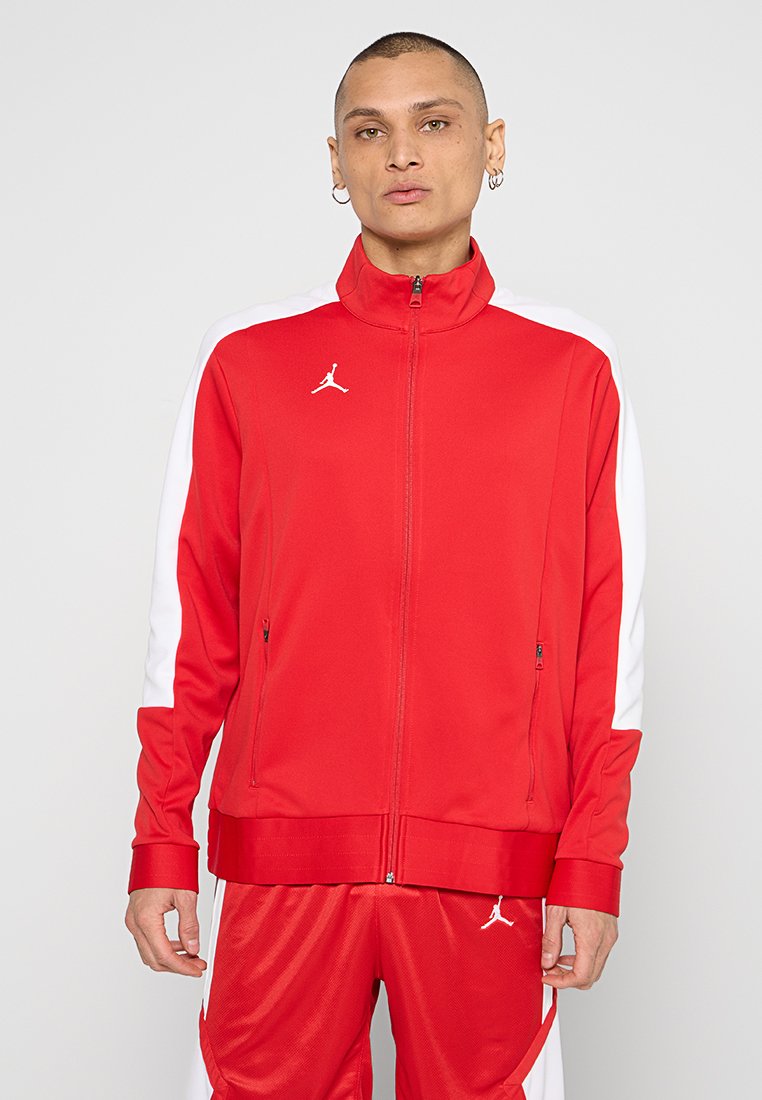 Chaqueta atlética roja hecha de tejido suave con paneles laterales blancos, cremallera de longitud completa y logo bordado en el pecho.