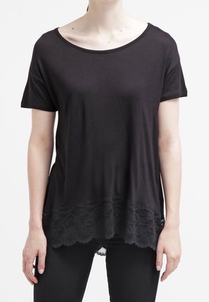 Donna che indossa un top nero a maniche corte con scollo a barca e pizzo lungo l'orlo, abbinato a pantaloni neri, in piedi davanti a uno sfondo bianco.
