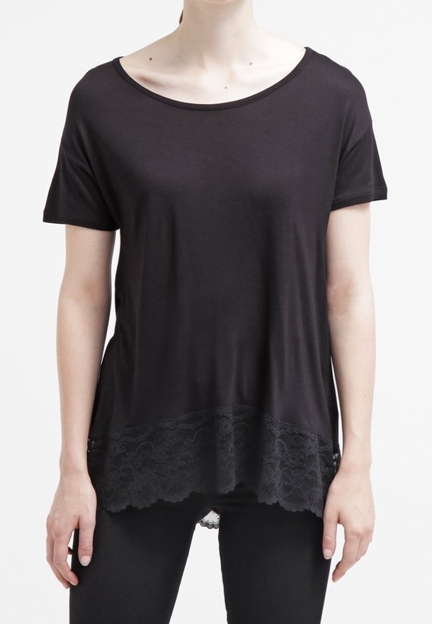 Kvinna som bär en kortärmad svart topp med rundad hals och spetsdetaljer vid nederkanten, kombinerad med svarta byxor, stående mot en vit bakgrund.