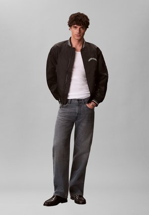 Jeune homme portant une veste aviateur noire Calvin Klein, un débardeur blanc, un jean gris et des chaussures habillées noires, debout les mains dans les poches.