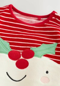 Punainen ja valkoinen raidallinen kangas, jossa on aplikoitu hymyilevä kasvot, vihreitä holly-lehtiä ja kolme punaista marjaa juhlavaatteissa.