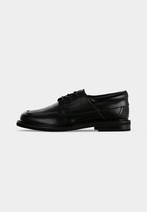 Chaussure habillée pour homme en cuir noir avec lacets, talon bas et nom de marque embossé sur le côté, vue de profil sur fond blanc.