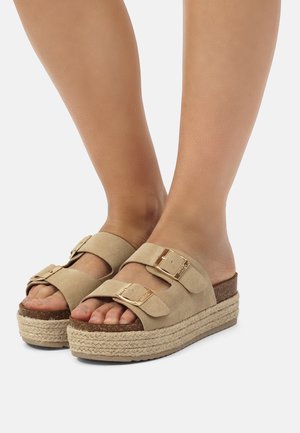 Füße mit beigen Plateausandalen mit zwei Schnallenriemen und geflochtenen Seilsohlen vor hellem Hintergrund.