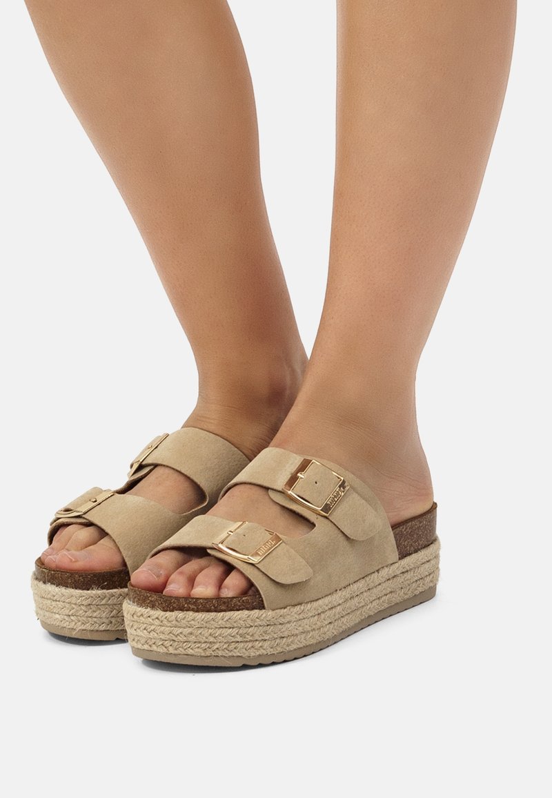 Pieds portant des sandales compensées beiges avec deux brides à boucle et des semelles en corde tressée sur un fond clair.