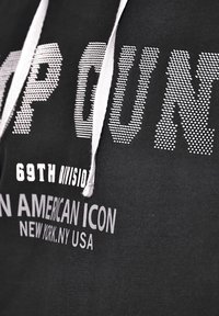 Svart huvtröja med vita dragsnören. Har stor tryckt text: "TOP GUN" i prickad stil, "69TH DIVISION" och "AMERICAN ICON" nedan.