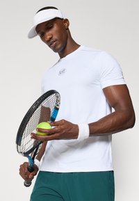 Valge sportlik t-särk ümmarguse kaelusega ja EA7 logo, koos roheliste lühikeste pükstega. Kinni hoides rohelise tenisepalli ja reketit, peas valge piil.