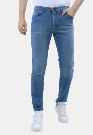 Man draagt slim-fit blauwe jeans met knoop- en ritssluiting, gecombineerd met witte sneakers, staat met één hand in de zak.