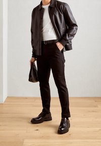 Chaqueta bomber de cuero negra sobre una camiseta blanca, combinada con pantalones cargo negros y zapatos con cordones. Sostiene una bolsa negra. Fondo de suelo de madera.